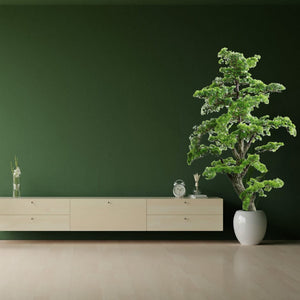 Künstlicher, detaillierter Bonsai mit weissem Topf vor einer grünen Wand mit Regal daneben
